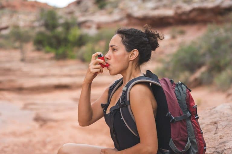 Woman with a backpack on in a desert setting is using a red inhaler. 